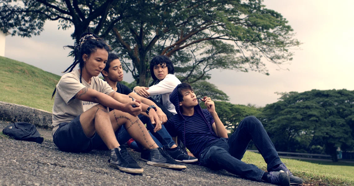 A group of four young people sit together on a grassy hill in a scene from the film Faeryville, surrounded by lush trees.