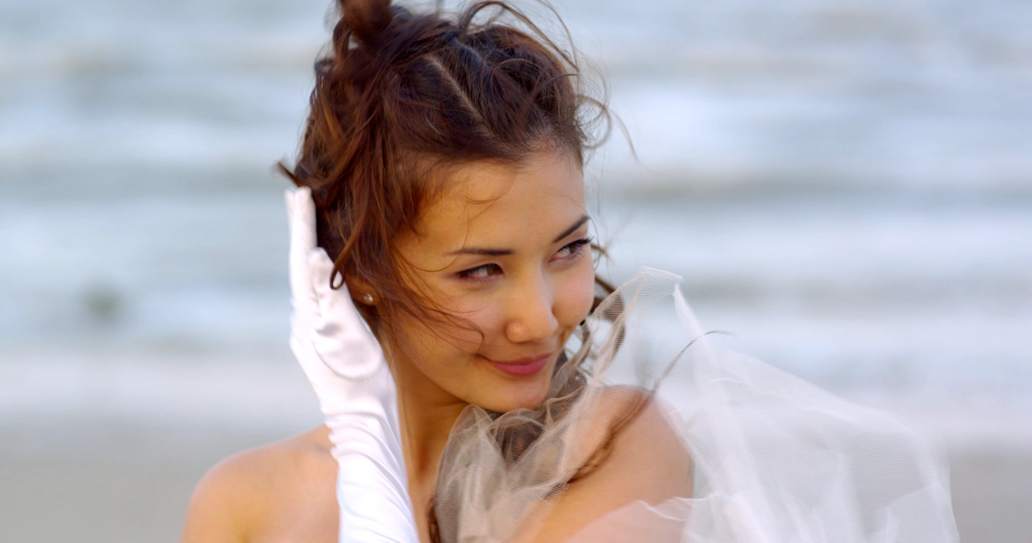 A still from the film "Forever" shows a woman in a white dress smiling on a beach, with wind-tousled hair and ocean in the background.