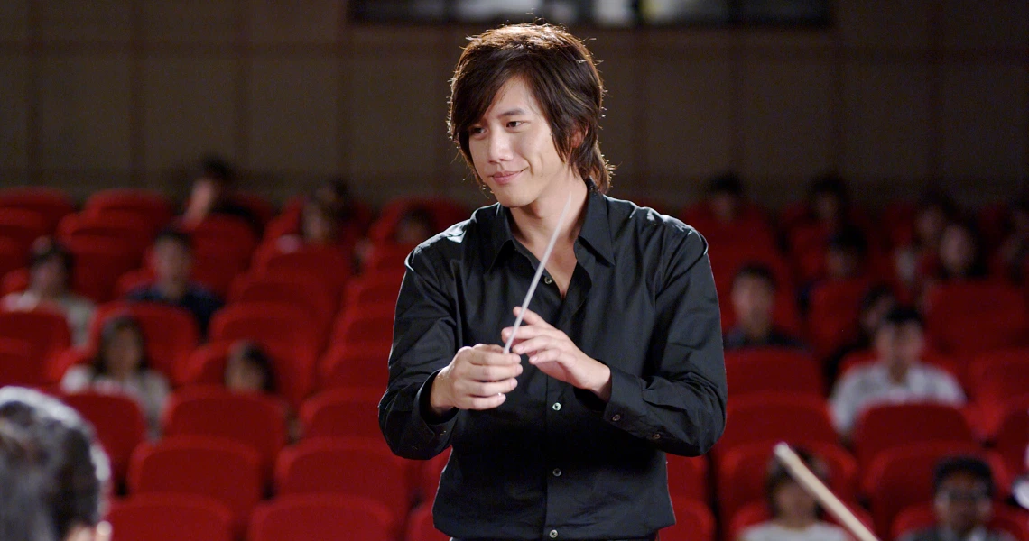 A young conductor in a black shirt smiles while holding a baton in an auditorium, a scene from the film "Forever".