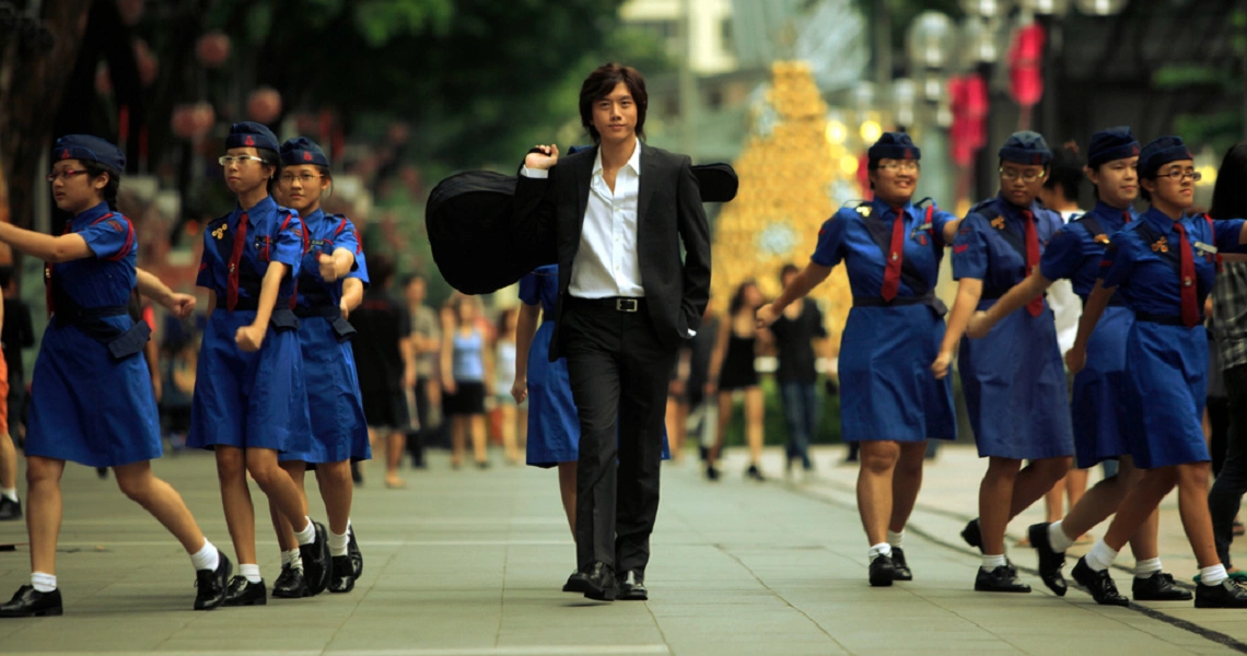 A scene from the film "Forever" shows a man in a suit carrying a guitar case, walking between two lines of uniformed schoolgirls on a street.