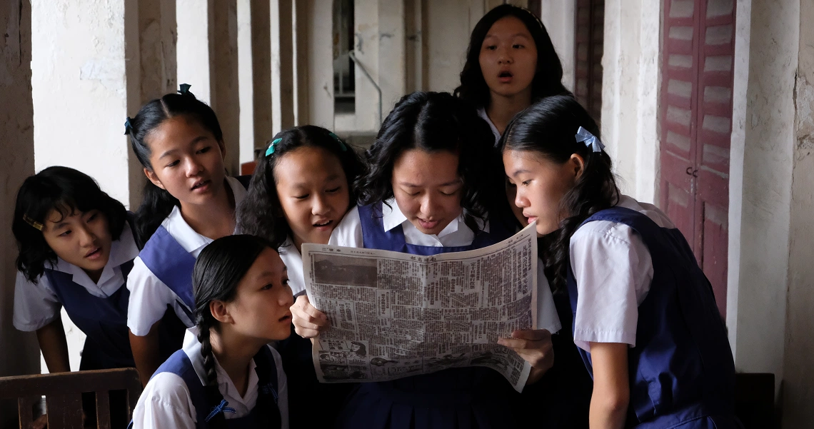 Students in school uniforms gather to read a newspaper in a scene from the film "From Victoria School To Ang Mo Kio".