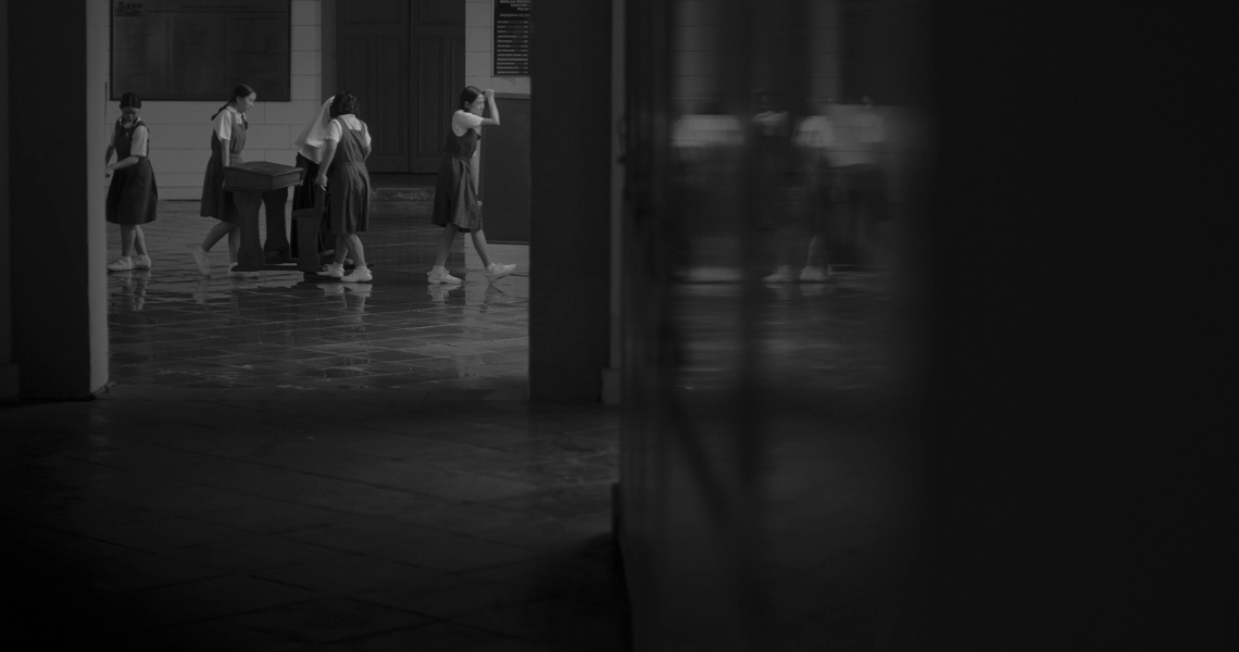 Atmospheric black and white film still from "From Victoria School To Ang Mo Kio" showing blurred figures in a shadowy corridor.