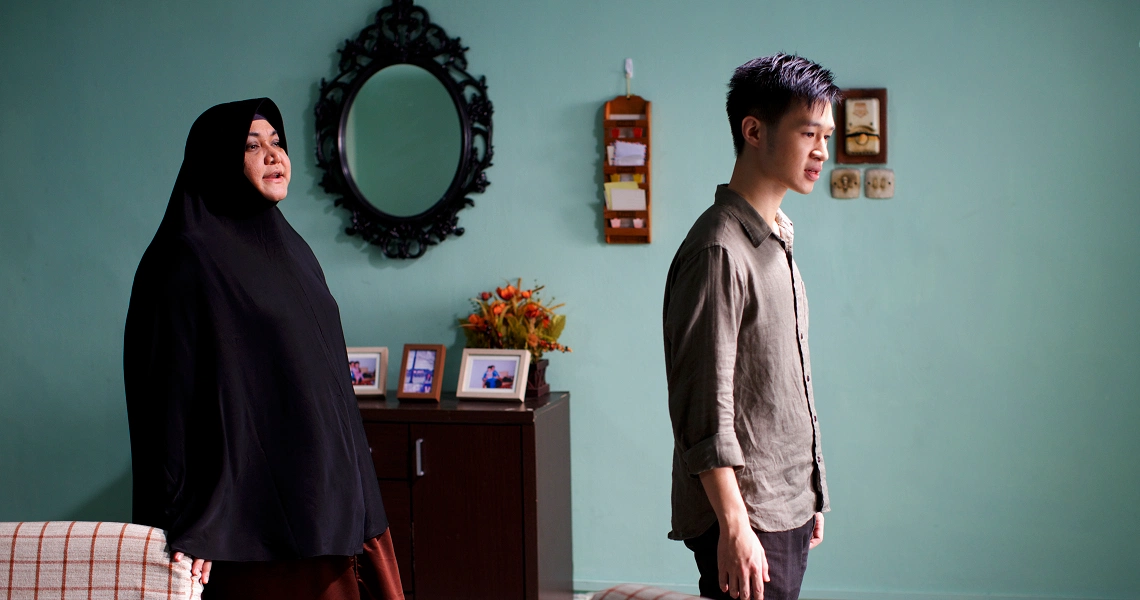 A scene from "Fundamentally Happy" shows a woman in black hijab and a man standing apart in a room with teal walls and ornate decor.