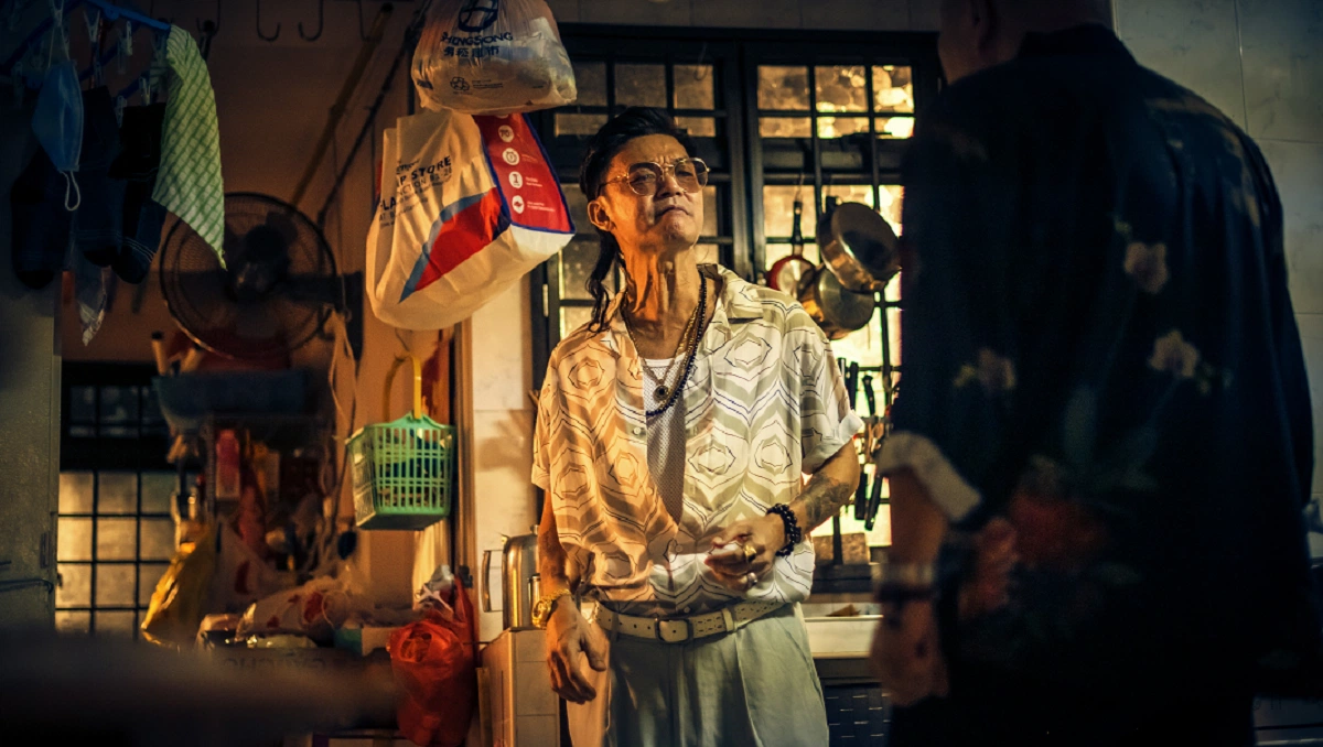 A scene from the film "Geylang" showing a character in a cluttered, dimly lit room filled with hanging clothes and various objects.