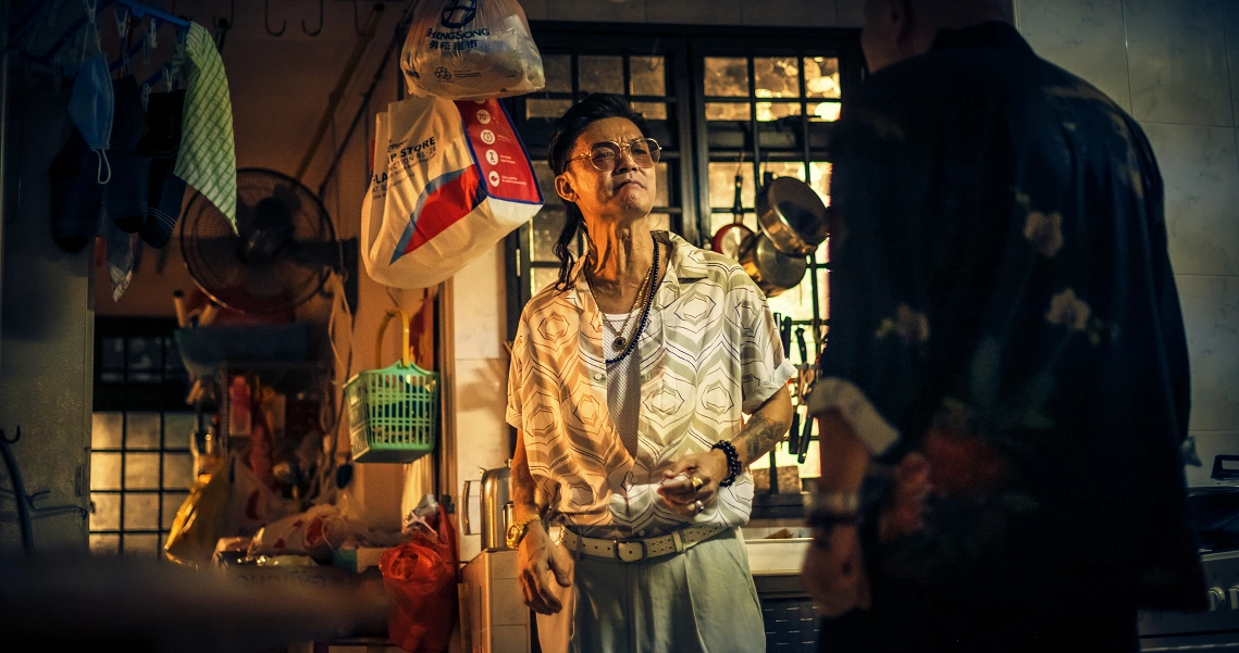 A scene from the film "Geylang" showing a character in a cluttered, dimly lit room filled with hanging clothes and various objects.