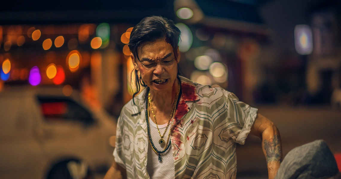 A man in a white patterned shirt with a serious expression walks through a colorful nighttime street scene in the film Geylang.