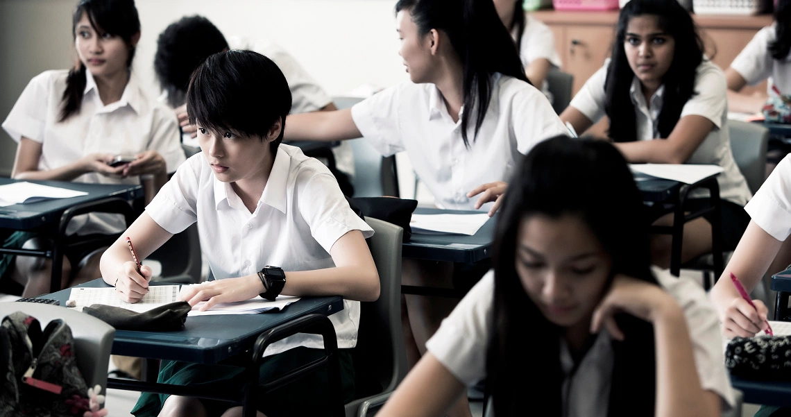 A classroom scene from the film Ghost Child shows students in white uniforms focused on their studies, capturing the school setting.