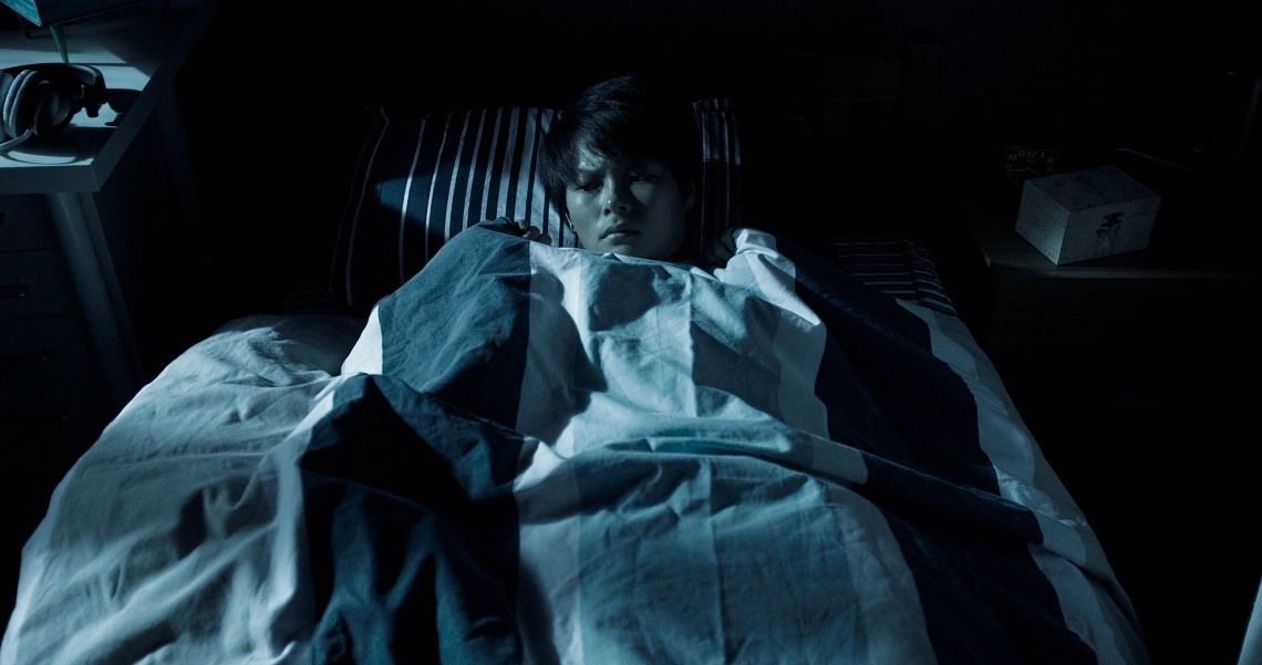 A tense scene from Ghost Child film shows a person lying in bed, covered in white sheets, in a dimly lit room, creating an eerie atmosphere.