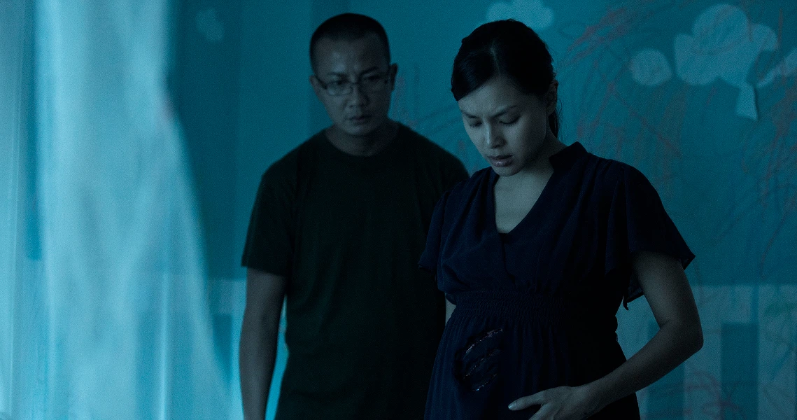 A tense scene from the film Ghost Child shows a pregnant woman in a dark room, with a concerned man standing behind her.