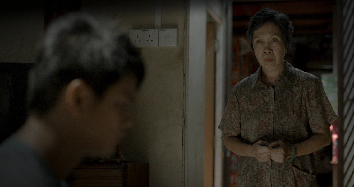 A tense scene from the film "Ghost On Air" showing an elderly woman in a dimly lit room, facing another person whose back is visible.