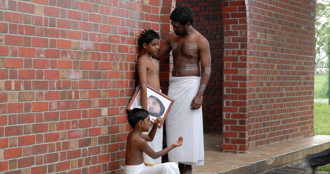 A poignant scene from Gurushetram shows three characters against a brick wall, one holding a framed portrait, capturing a moment of reflection.