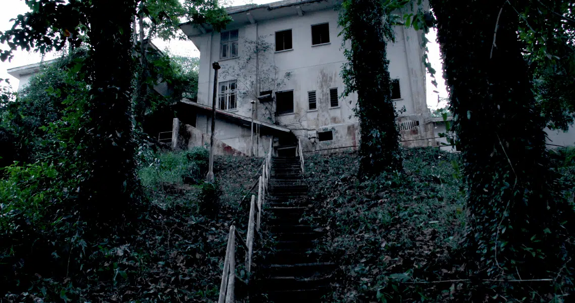 Eerie film still from "Haunted Changi" showing an abandoned building with overgrown stairs leading to a dilapidated white structure.