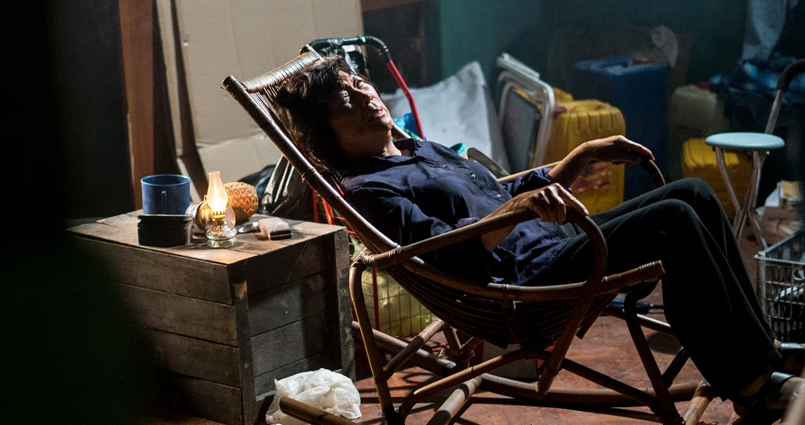 A person relaxes in a wooden chair, surrounded by clutter in a dimly lit room, in a film still from "Hell Hole".