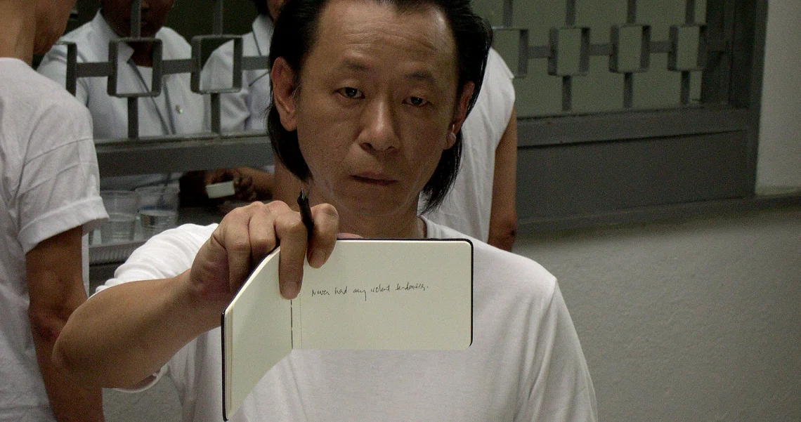 A man in a white shirt holds up a small piece of paper with writing on it in a scene from the film "Here".