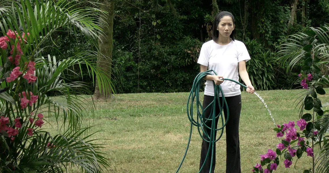 A woman in a white shirt waters plants in a lush garden setting for the film "Here", showcasing a scene from the movie.