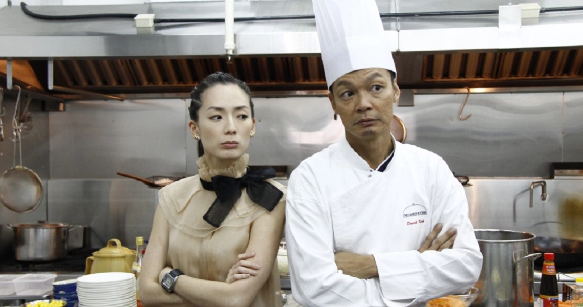 A scene from the film Homecoming shows a woman in a beige dress and a chef in white uniform standing in a restaurant kitchen.