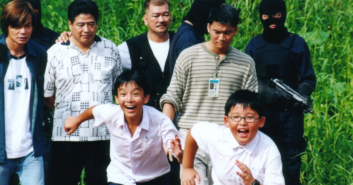 A film still from "I Not Stupid" showing a group of young boys and men posing outdoors, with one masked figure in the background.