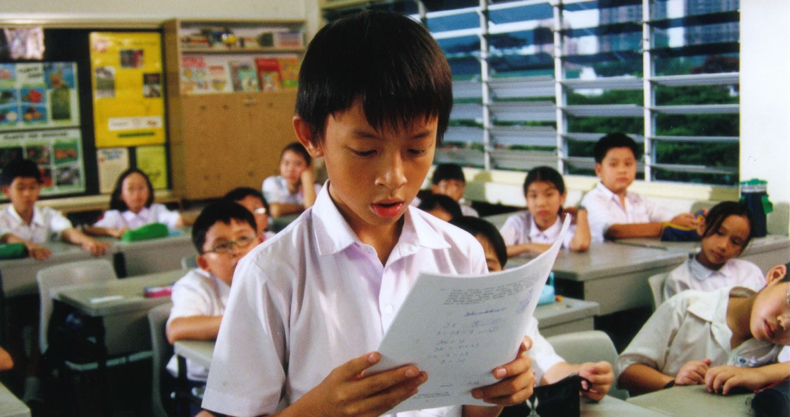 A scene from the film "I Not Stupid" showing a young student reading aloud in a classroom full of attentive classmates.