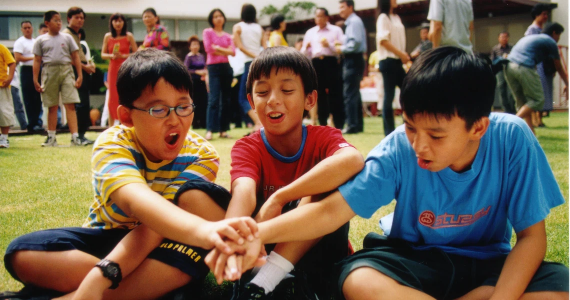 Three young boys from the film "I Not Stupid" sit on grass, laughing and playing together in a school setting.