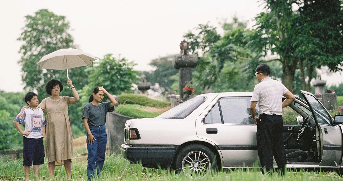 A family stands near a car in a rural setting, a scene from the Singaporean film Ilo Ilo.