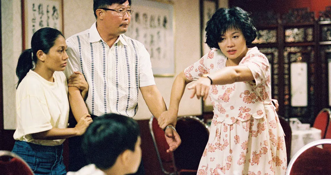 A scene from the film Ilo Ilo showing a family in a living room, with a woman in a floral dress gesturing animatedly.