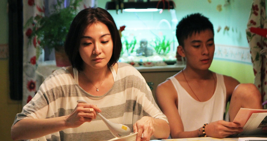 A scene from the film "Imperfect" showing two young people sitting at a table, one focused on a task while the other looks on.