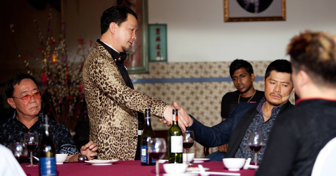 A scene from the film "Imperfect" shows a group dining at a table while a man in a leopard-print jacket pours drinks.