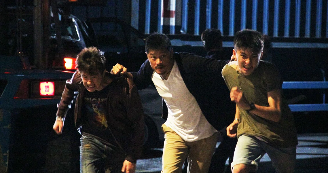 A still from the film "Imperfect" showing three young men running urgently at night, illuminated by dim lighting.