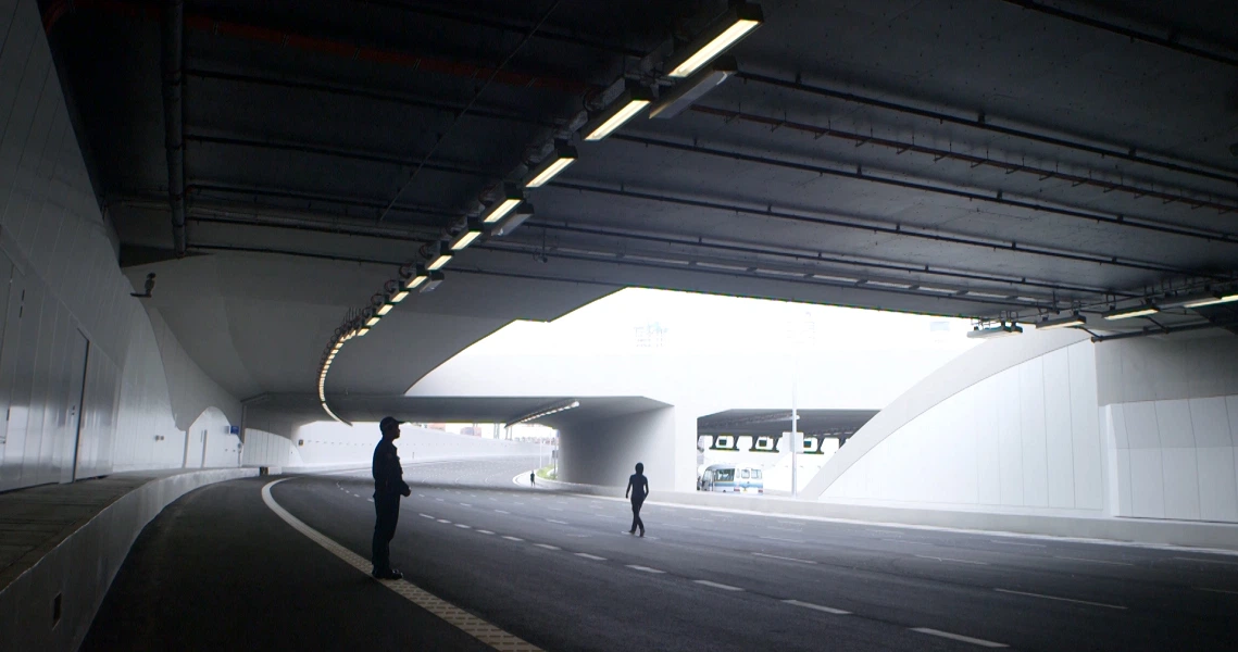 A scene from "In Time to Come" showing silhouettes of people walking through a modern, minimalist underground passage with stark lighting.