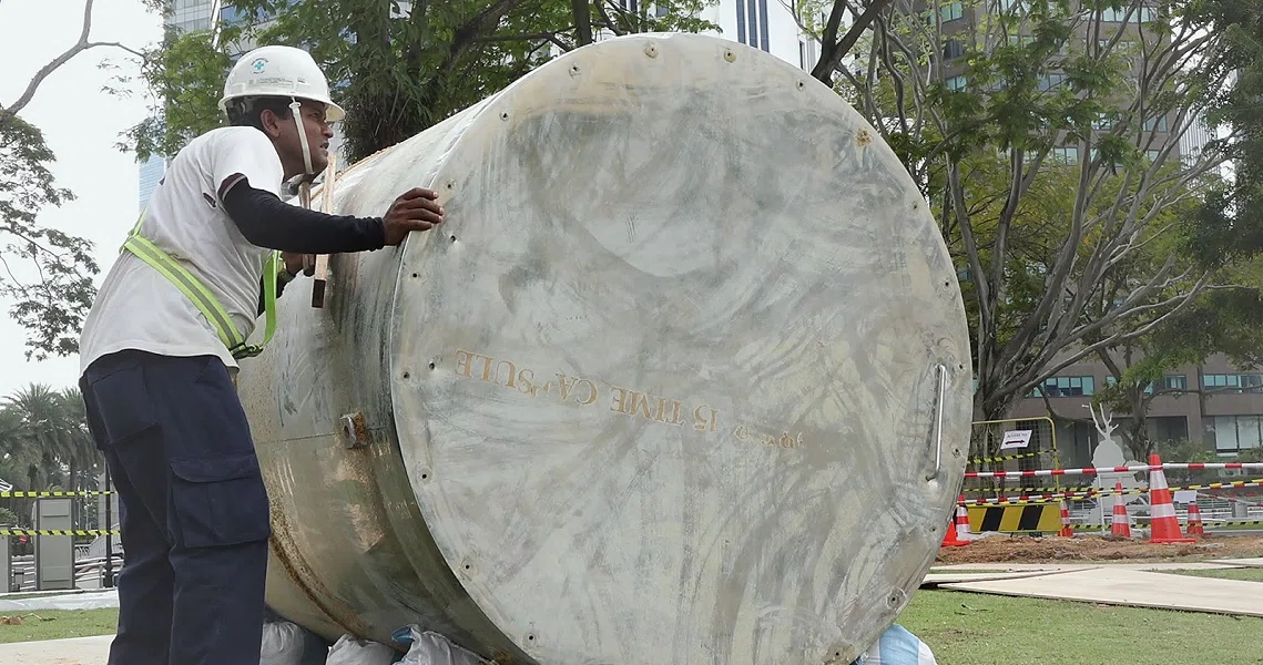 A construction worker in safety gear examines a large time capsule in an urban setting, a scene from the film "In Time to Come".