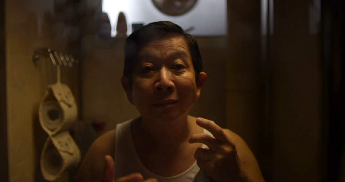 A man in a white tank top stands in a dimly lit bathroom, pointing at himself in the film still from "Judgement Day".