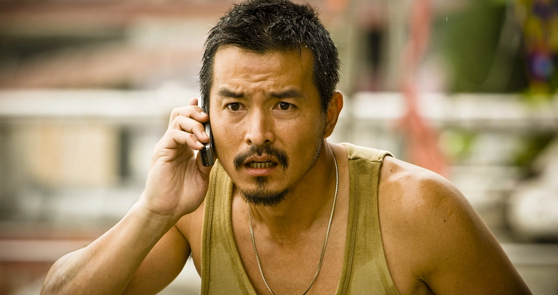 A tense scene from the film Kidnapper shows a man in a tank top speaking on a mobile phone with a concerned expression.