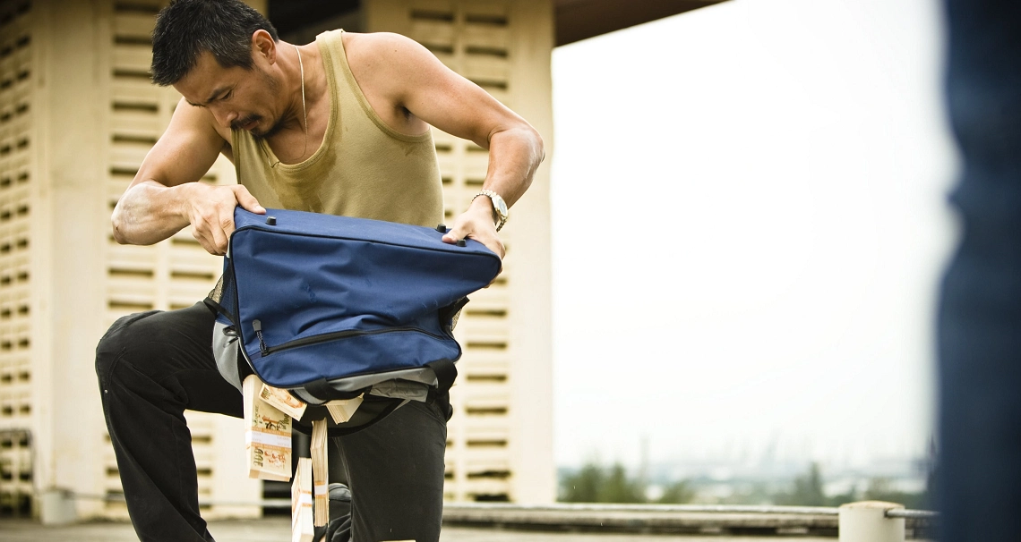 A shirtless man in the film "Kidnapper" searches through a blue bag, creating tension in this dramatic scene.