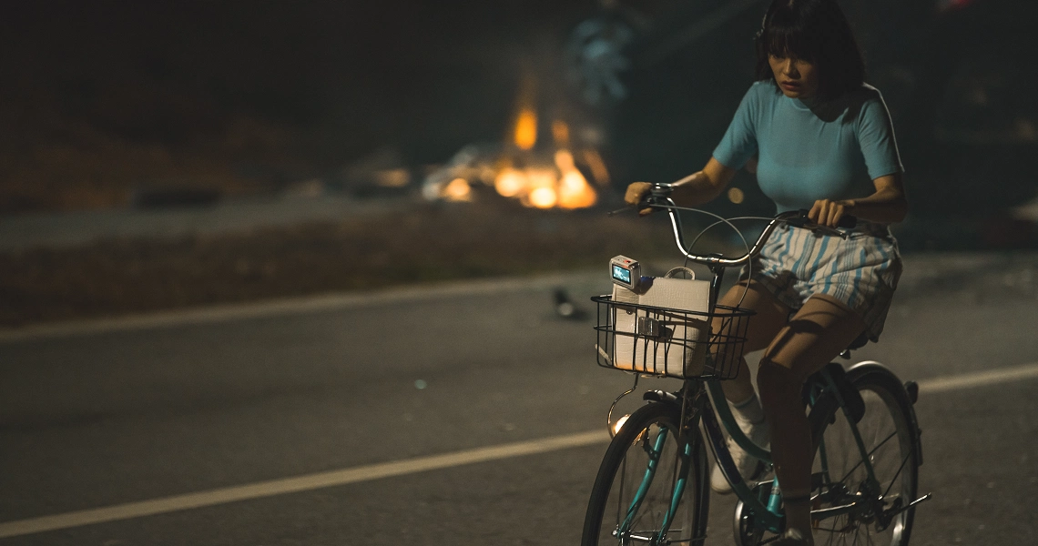 A woman rides a bicycle at night in a scene from the film "Late Night Ride", with a fire visible in the background.