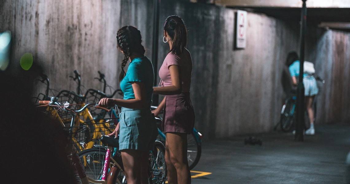 Film still from "Late Night Ride" showing two women standing next to a bicycle in a dimly lit urban setting.