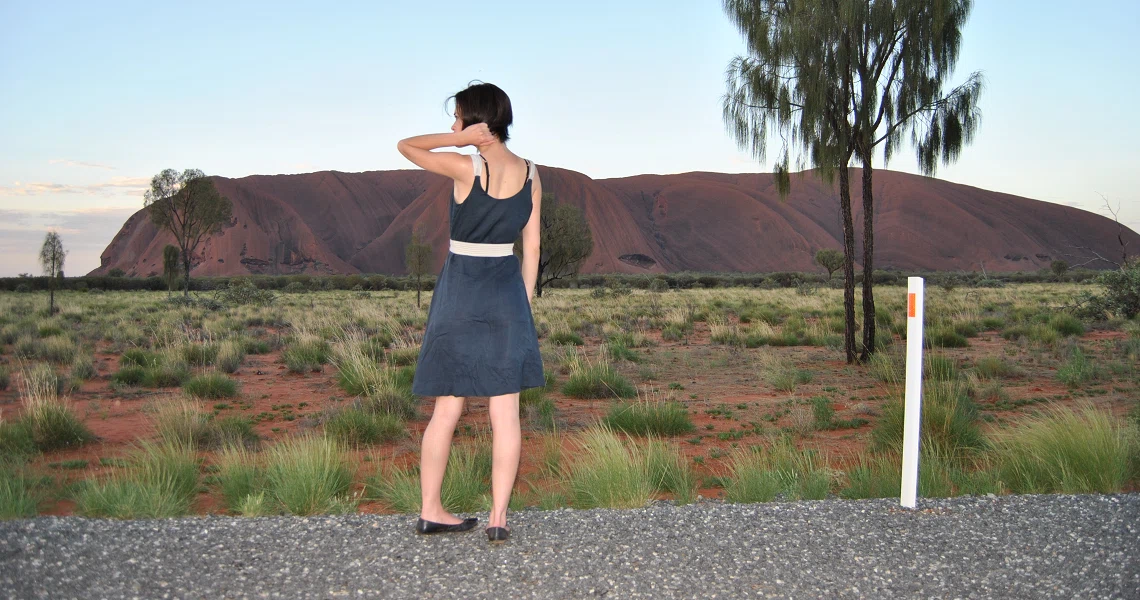 A film still from Liberta showing a woman in a blue dress standing on a road, facing a large red rock formation in a desert landscape.