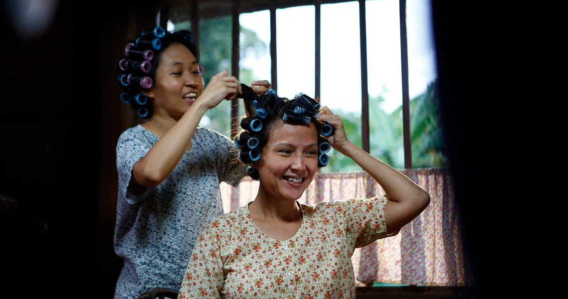 A scene from "A Long Long Time Ago" showing two women smiling, one styling the other's hair in a vintage setting.