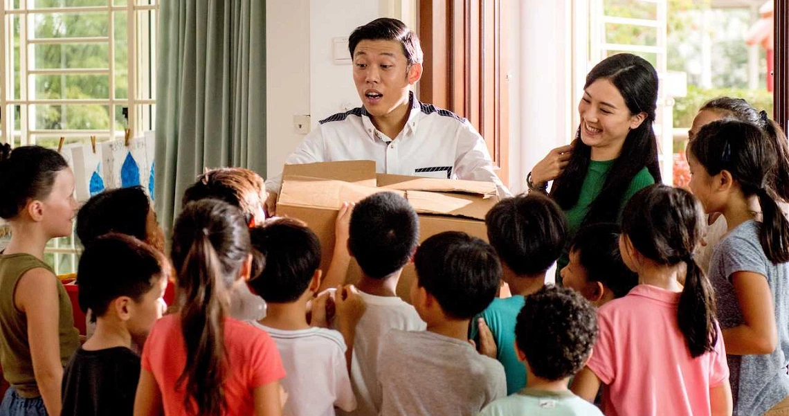 A scene from the film Lucky Boy shows two adults surrounded by a group of children in a bright, indoor setting.