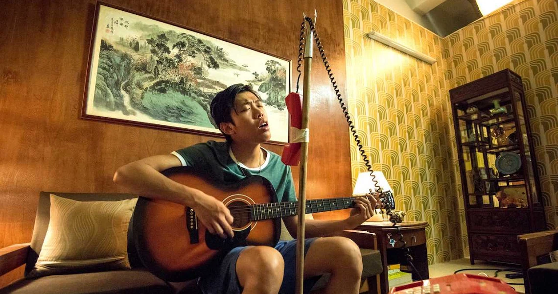 A scene from the film Lucky Boy shows a young man playing an acoustic guitar in a cozy room with Asian-inspired decor.