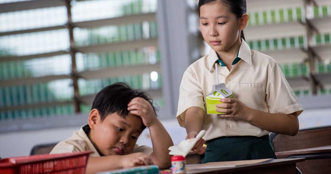 A scene from the film Lucky Boy showing a schoolgirl holding a drink while a tired boy rests his head on a table.