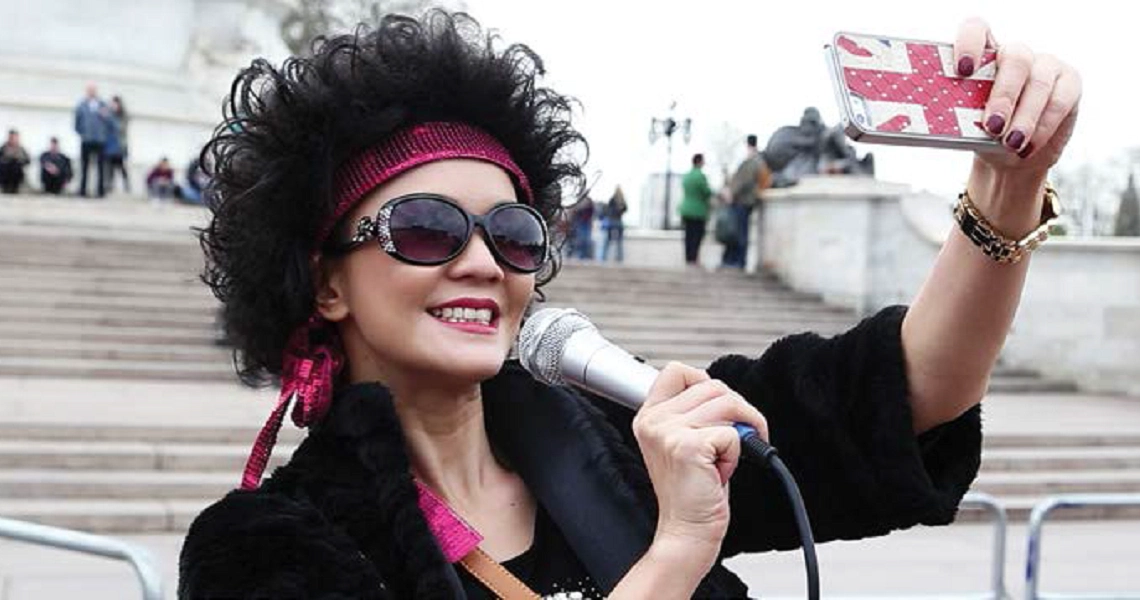 A woman with curly hair and sunglasses holds a microphone and Union Jack flag in a scene from the film "Lulu The Movie".