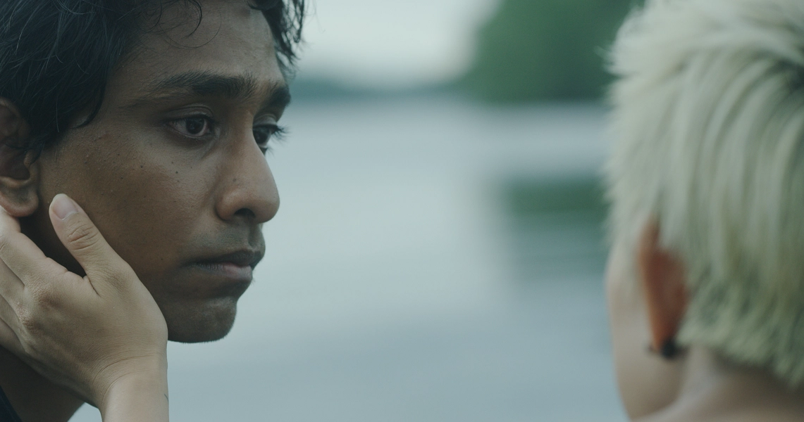 A film still from "Malam" showing a close-up of a person's face with another person's hand touching their cheek.