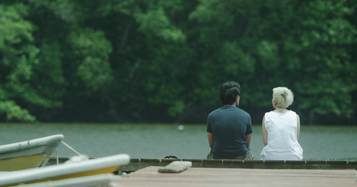 A still from the film Malam shows two people sitting on a bench by a lake, surrounded by lush greenery, creating a serene atmosphere.