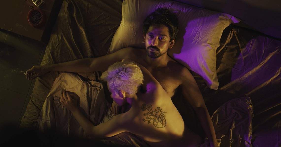 A film still from "Malam" shows a shirtless man with tattoos lying in bed, bathed in purple light, creating a moody atmosphere.