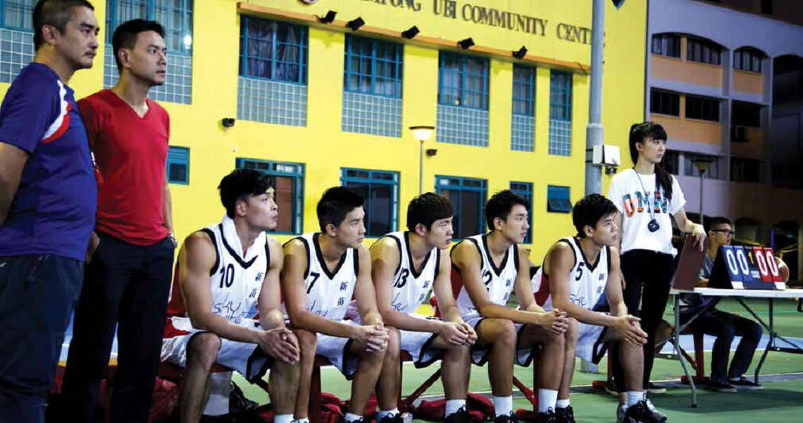 Film still from "Meeting The Giant" showing a basketball team sitting on bench outside yellow community center building.