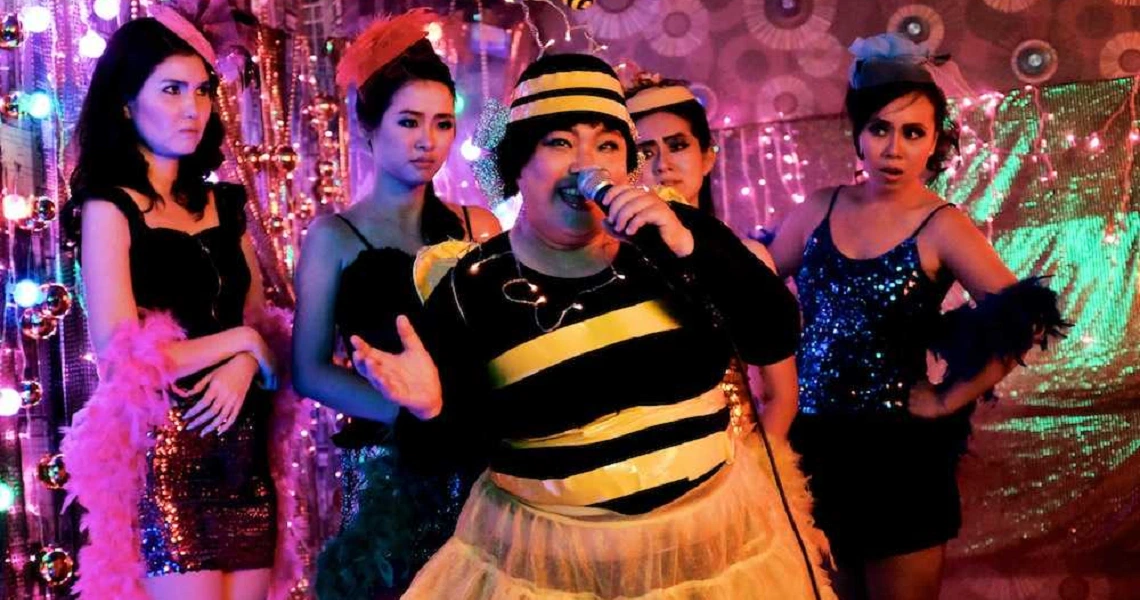 A colorful scene from the film "Mr Unbelievable" showing a person in a bee costume surrounded by four women in dark outfits.