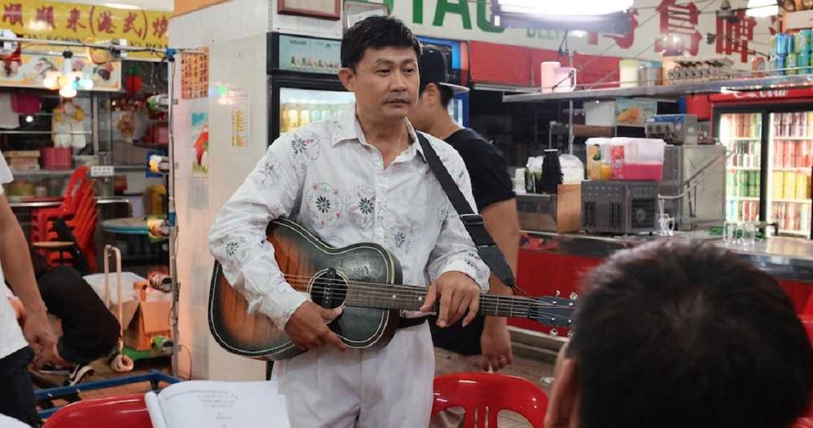 A scene from the film "Mr Unbelievable" showing a man in white clothing holding a guitar in a colorful outdoor market setting.