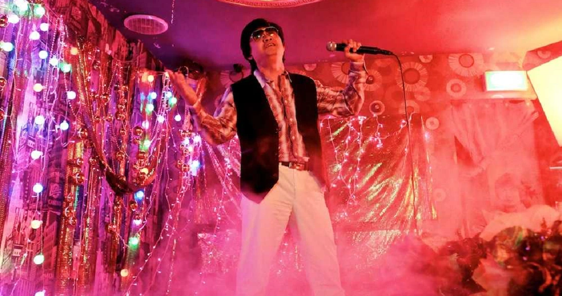 A vibrant scene from "Mr Unbelievable" shows a performer singing on stage amidst colorful lights and shimmering decorations.