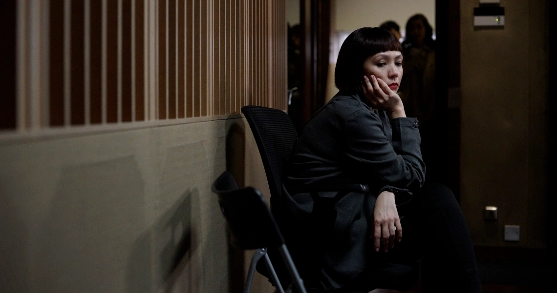 A pensive woman in dark clothing sits alone in a dimly lit hallway, contemplating her choice in a scene from "Ms J Contemplates Her Choice".