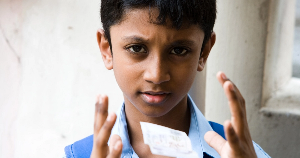 A boy in a blue shirt and backpack holding a white paper, performing a magic trick.